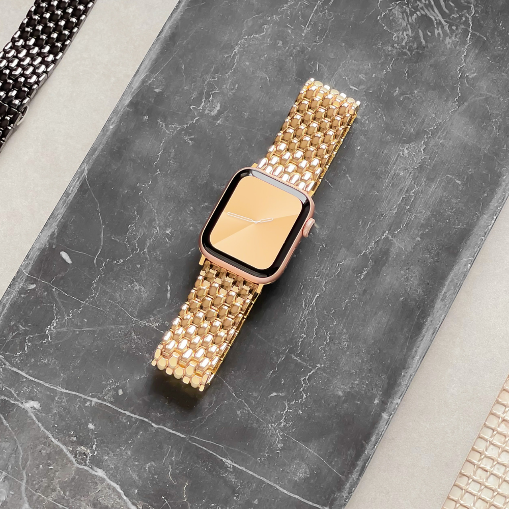 A smart watch with Coconut Lane’s Mermaid Apple Watch Strap - Gold, featuring a pink face, is displayed on a grey marble surface. The background contains partial views of similar watch bands, including a Zinc Alloy Adjustable Strap, placed on either side of the marble surface.